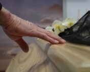 Elderly hand resting on a wooden casket during a funeral