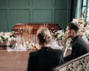 Man and woman sitting near decorated wooden casket during funeral service